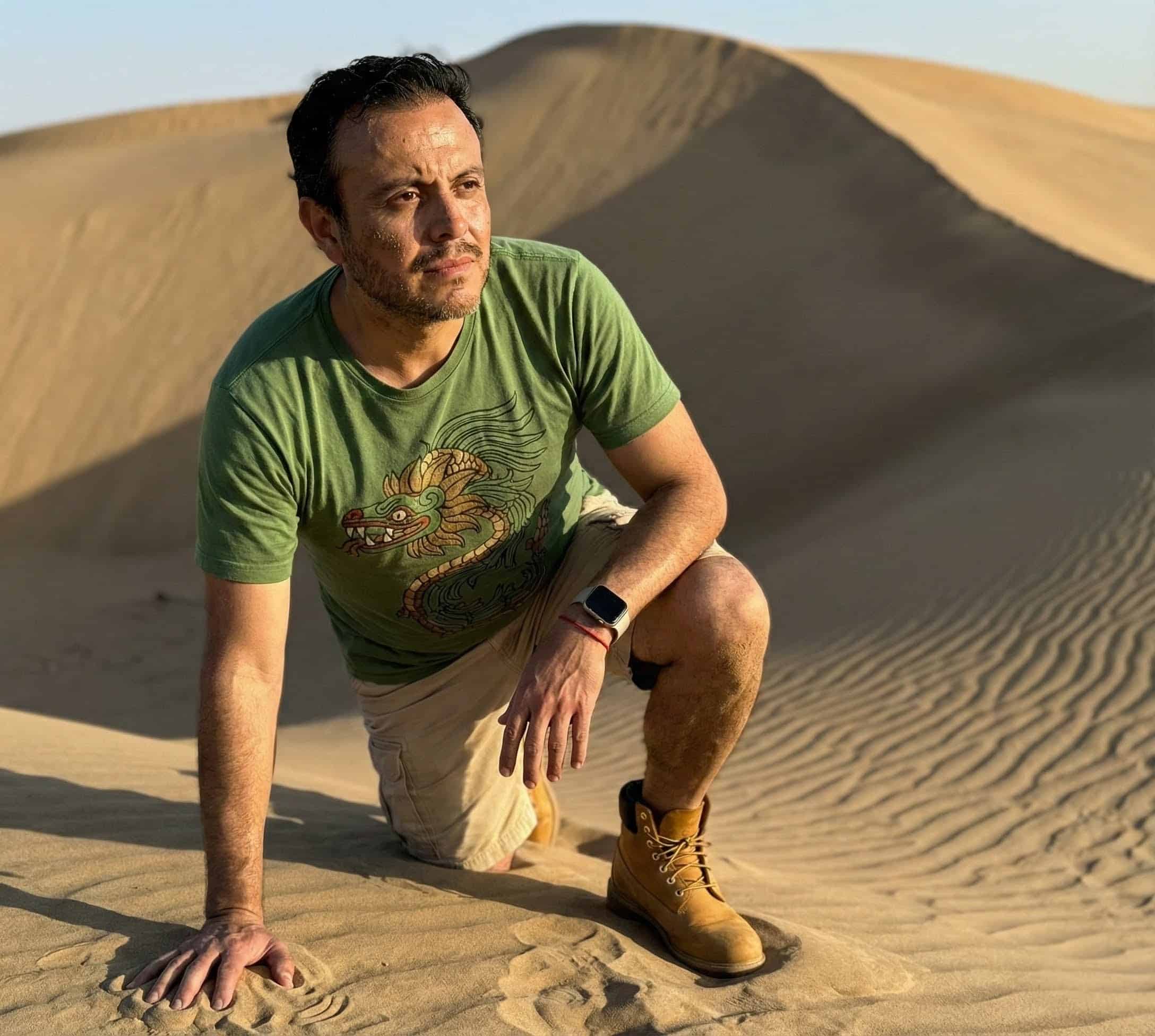 Juan Pablo García fija su mirada en un punto, su rostro está lleno de arena y su brazo derecho sostiene el suelo arenoso mientras que su brazo izquierdo descansa en su rodilla de ese mismo lado. Está a punto de incorporarse para comenzar a andar y salir de su desierto personal.